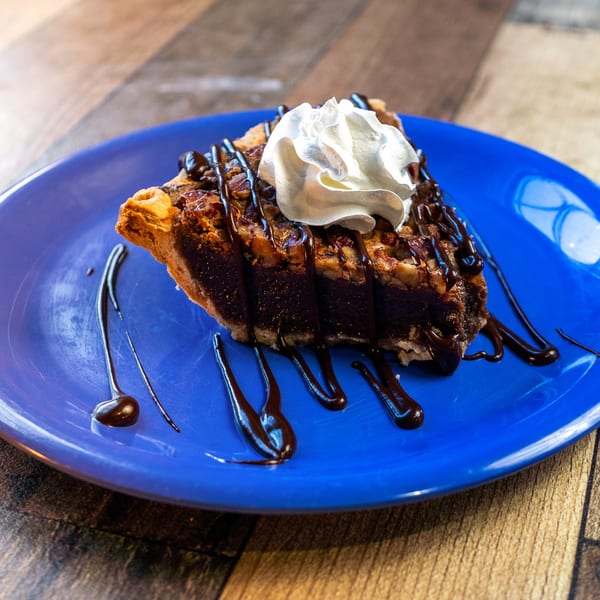 A slice of pecan pie with chocolate drizzle and whipped cream on a blue plate.